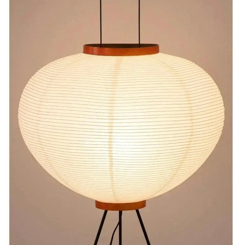 A glowing spherical paper lantern-style pendant lamp with horizontal ribbing, suspended by black cords with a warm brown top and bottom trim, set against a plain beige background.