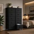 A tall black fabric dresser with multiple drawers stands beside a bed in a styled bedroom. One lower drawer is open, showing folded bedding, and the scene includes a plant, framed wall art, a lamp, and a rug.