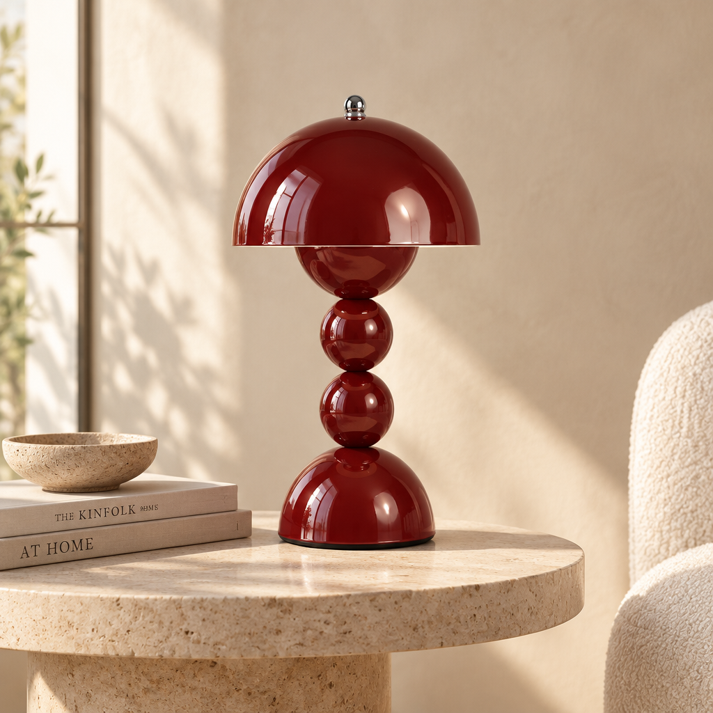 A glossy deep red table lamp with a dome-shaped shade and stacked spherical base, styled on a round stone side table in a warm, sunlit room.