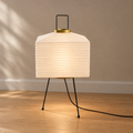 A modern table lamp with a pleated white paper shade, black metal tripod legs, and gold accents, shown lit in a minimalist room setting with a beige wall and light wood floor.
