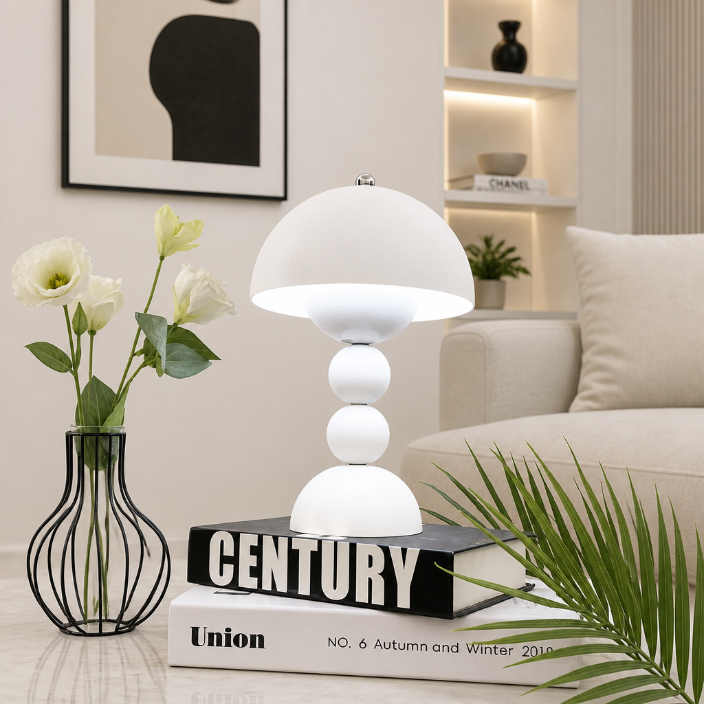 A modern white table lamp with a dome-shaped shade and three stacked spherical bases sits on books in a styled living room setting. The lamp is surrounded by decorative props including a black wire vase with white flowers, a green palm leaf, and a beige sofa in the background.