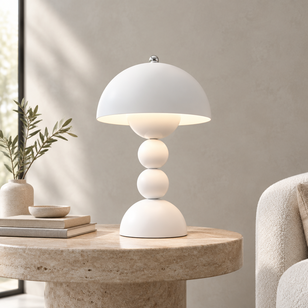 A white mushroom-shaped table lamp with a domed shade and stacked spherical base, lit from within, placed on a round stone side table in a softly styled interior setting.