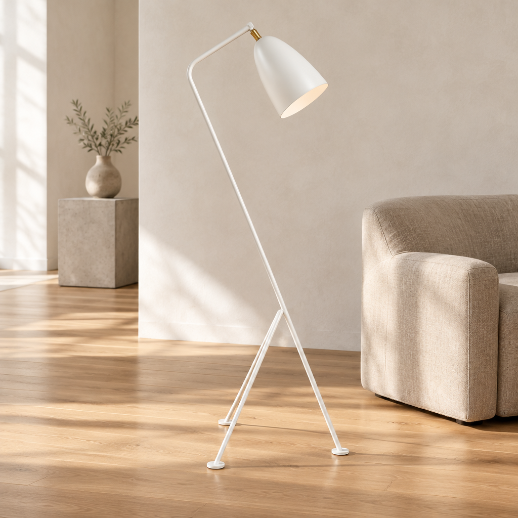 A modern white floor lamp with a tripod base and adjustable curved arm stands on a light wood floor next to a beige armchair in a minimalist living room.