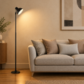 A modern black floor lamp with a conical shade stands lit next to a light beige sofa in a warm, contemporary living room setting.