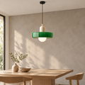 A modern pendant light with a glossy green circular shade, a cream cylindrical accent, and a glowing white globe bulb, hanging over a wooden dining table in a minimalist room with a textured beige wall.