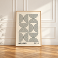 A framed Bauhaus 1919 art print with a black geometric line pattern on an off-white background, leaning against a white paneled wall on a light wood floor.