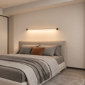 A modern minimalist bedroom featuring a low-profile horizontal wall light mounted above a bed with grey bedding and a rust-orange accent pillow, set against a warm beige wall.