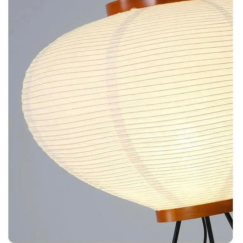 A close-up of a modern floor lamp with a large, ribbed, teardrop-shaped white shade that glows with warm light, framed by a brown wooden top and bottom trim with black metal legs visible at the bottom right.
