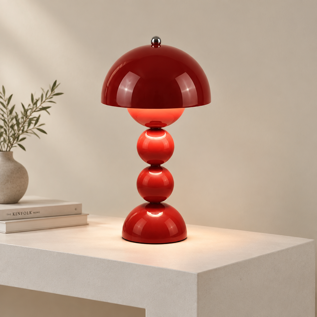 A glossy red table lamp with a mushroom-shaped dome shade and three stacked spherical bases, shown lit on a white pedestal against a warm beige wall. A small vase with greenery and stacked books sit to the left.
