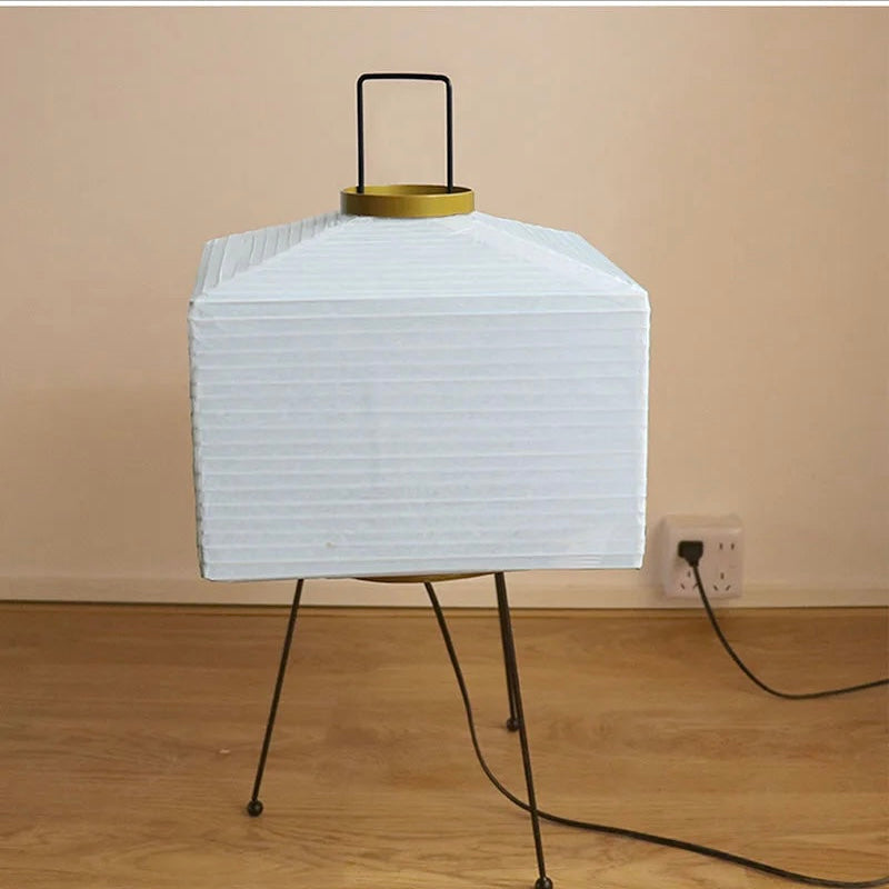 A modern table lamp with a white pleated rectangular shade, black metal tripod legs, and a gold-toned top collar, shown on a wooden floor against a beige wall.