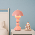 A retro mushroom-shaped table lamp in a dusty pink color, glowing with a warm light, styled on a white bedside table next to a wooden decorative tree against a light blue wall.