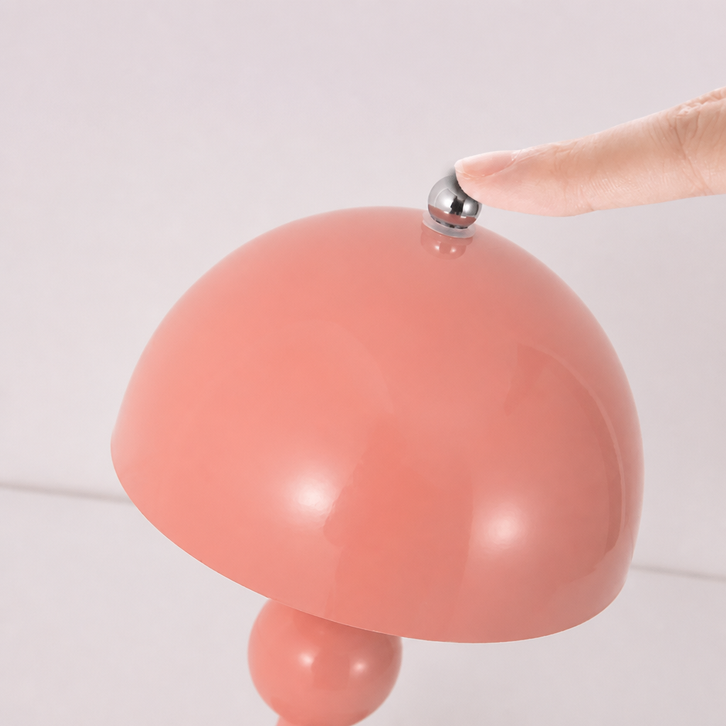 A close-up of a coral pink mushroom-shaped table lamp with a glossy finish, featuring a finger pressing a small silver metal button on top of the dome shade.