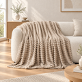A chunky knit throw blanket in warm beige is draped over a cream-coloured sofa in a bright, modern living room.