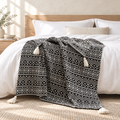 A black-and-off-white patterned throw blanket with cream tassels draped over a bed with white bedding in a bright, modern bedroom.