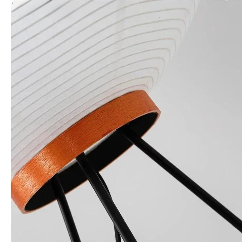 Close-up of a modern floor lamp with a white ribbed paper lantern shade, a warm copper-toned metal band, and slim black metal legs on a light gray background.
