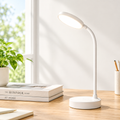 A white adjustable desk lamp with a circular base and curved neck stands on a wooden desk beside a potted plant and books in a bright indoor setting.