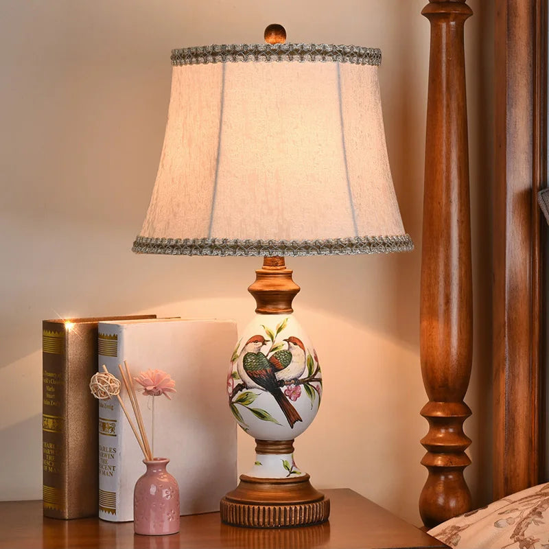 Bird-pattern resin table lamp with chenille shade displayed beside books and a small pink vase