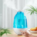 Blue and white humidifier on a table with a hand adjusting it, fruits in the background.