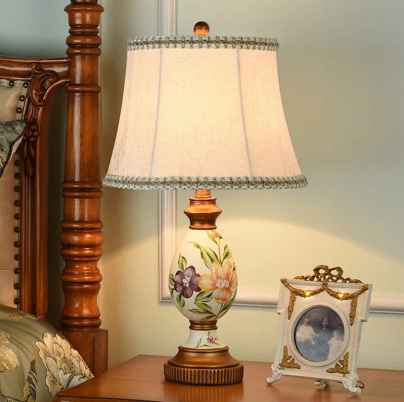 Hand-painted floral resin table lamp on a wooden nightstand beside an antique-style picture frame