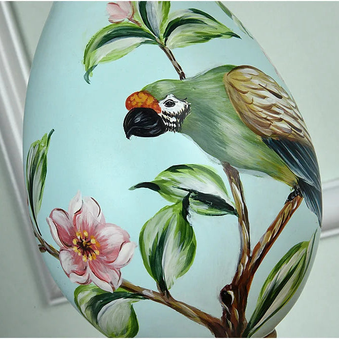 Resin table lamp with green parrot design and floral-print lampshade beside a framed bird illustration