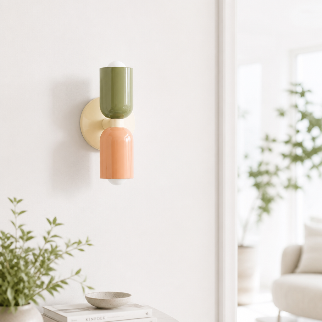 A modern two-light wall sconce mounted on a white wall in a bright, minimalist room. The fixture features two cylindrical shades stacked vertically: the top shade is olive green, the bottom shade is a warm peachy orange, and the wall mount is a soft cream/beige color.