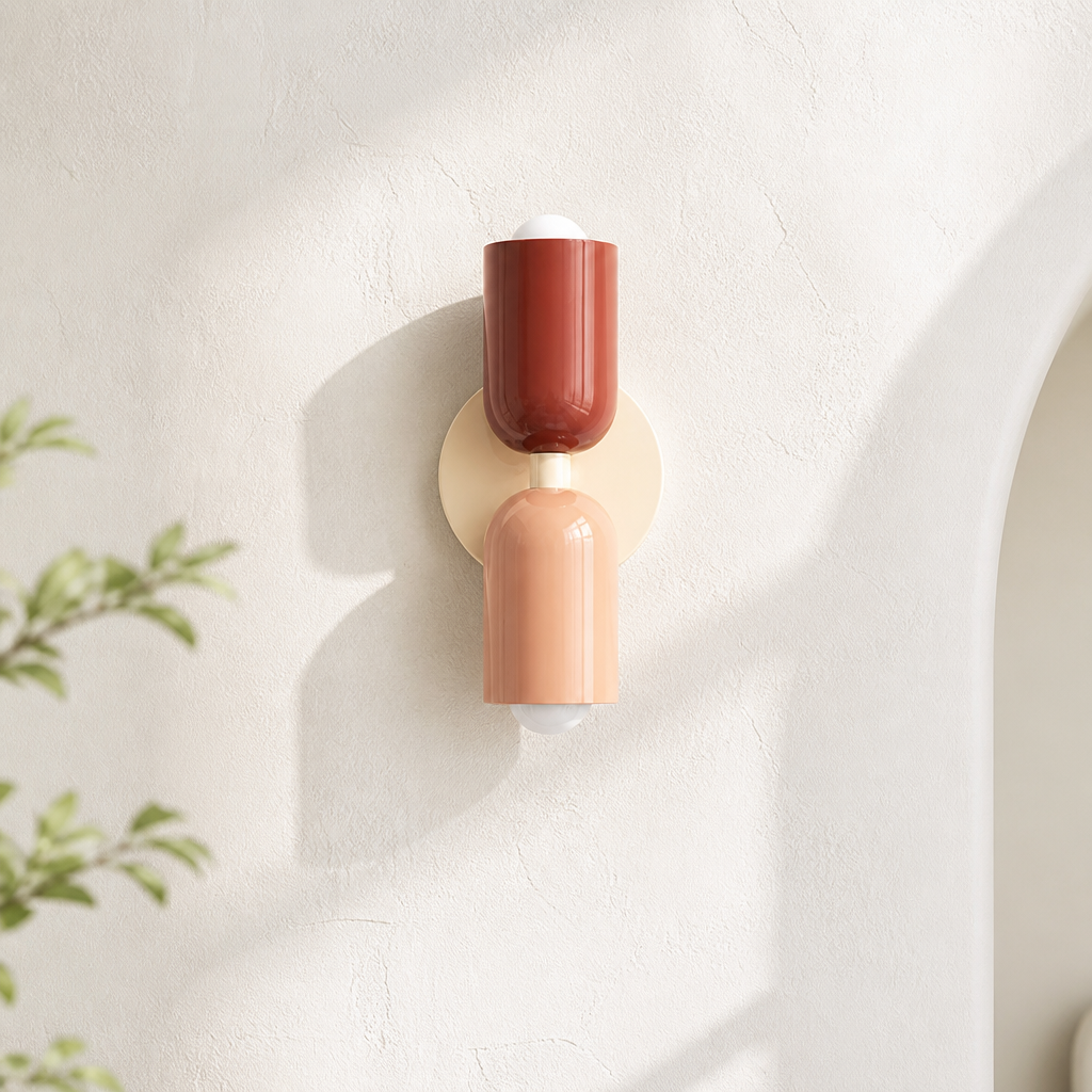 A modern wall sconce light fixture with two cylindrical shades in terracotta and peach, mounted on a round beige backplate against a textured off-white wall.