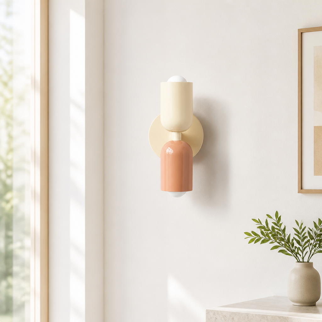 A modern wall sconce light fixture with a cream-colored upper cylinder, a terracotta-pink lower cylinder, and a beige circular backplate, mounted on a warm off-white wall in a styled interior setting.