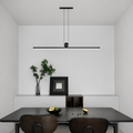 A minimalist modern linear pendant light in matte black hangs above a dark dining table with two wooden chairs in a clean, contemporary dining room setting.