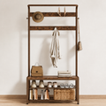 A rustic dark brown entryway coat rack with a bench, styled with a straw hat, beige coat, handbag, shoes, and a woven basket on a light wood floor against a white wall.