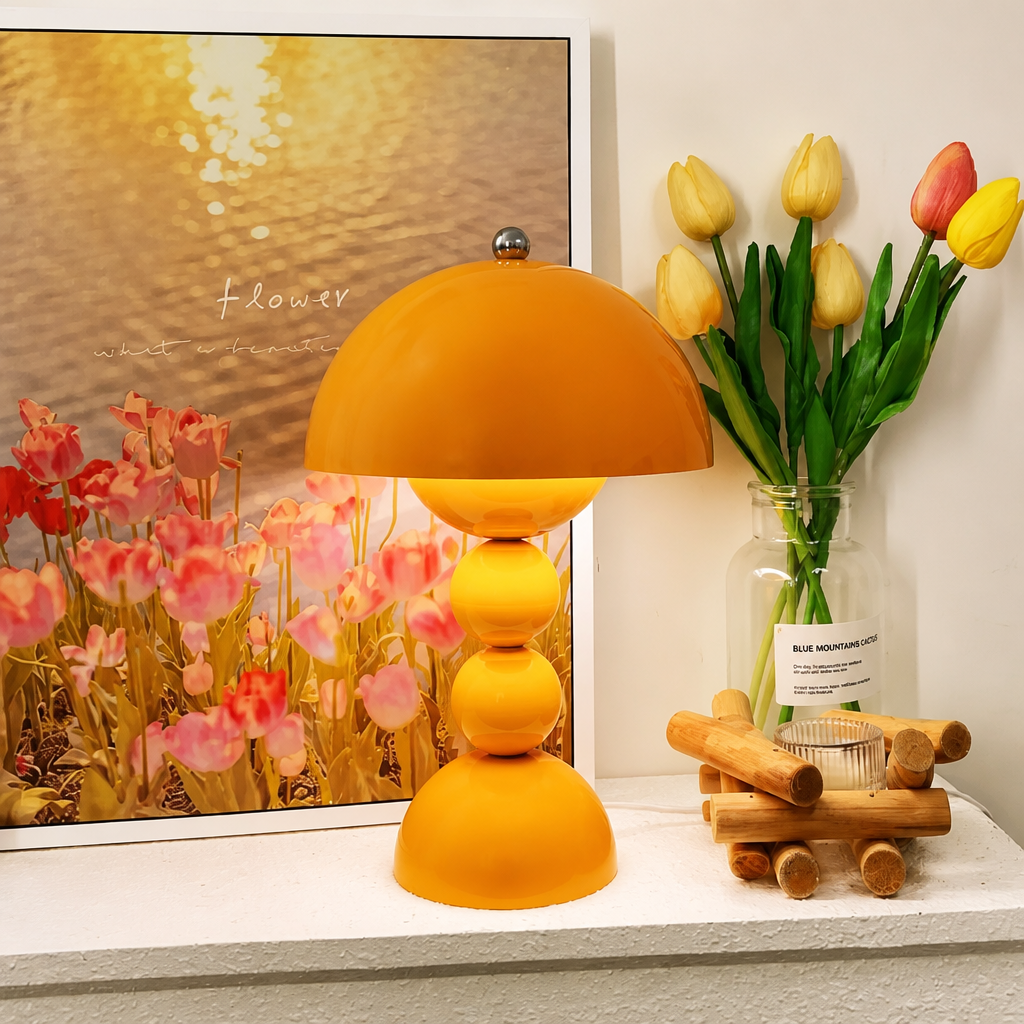 A mushroom-shaped table lamp with a warm yellow gradient shade and stacked spherical base, styled on a white shelf next to a framed tulip print, a glass vase with yellow tulips, and a small wooden toy boat.