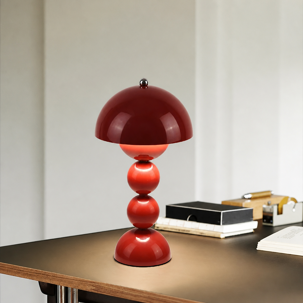 A glossy red mushroom-shaped table lamp with a domed shade and three stacked spherical bases, shown on a dark tabletop in an indoor setting with books and stationery blurred in the background.