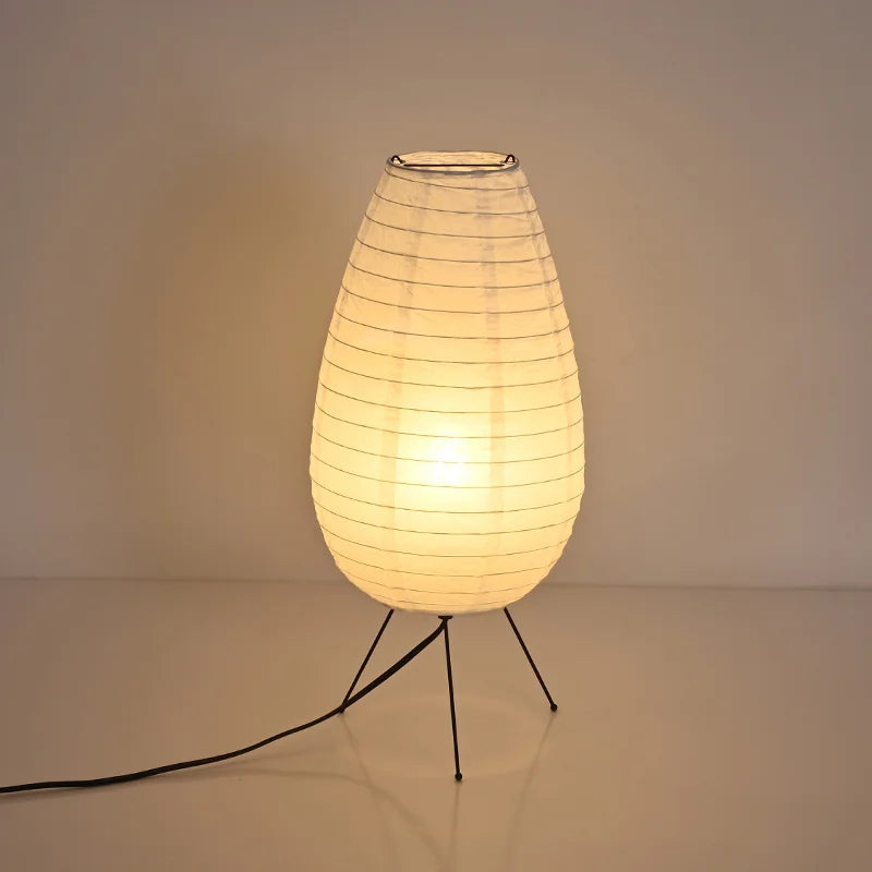 A table lamp with a glowing warm white ribbed teardrop-shaped shade on thin black tripod legs, set against a plain beige background.