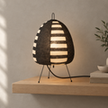 A woven egg-shaped table lamp with black and natural wood horizontal slats, glowing warm white when lit. It sits on thin black metal legs on a light wood floating shelf against a textured beige wall.