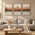 A three-piece framed wall art set depicting a serene mountain and lake sunset scene, styled above a light gray sofa in a warm, modern living room.