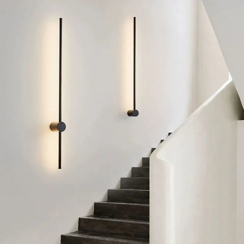 Modern minimalist wall sconces with slim black vertical rods and circular gold and dark metal accents, mounted on a white wall above a dark concrete staircase.