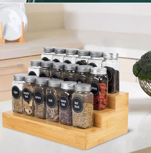 A collection of 12 glass spice jars with silver lids organized on a three-tier bamboo spice rack.