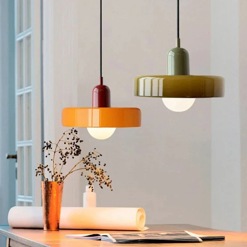 Two modern pendant light fixtures hang over a table in a bright interior. One has a glossy orange shade and a dark red stem, and the other has an olive green shade and a muted sage green stem.