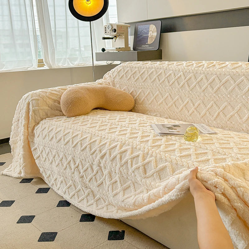 Beige textured blanket draped over a sofa with a hand holding it, in a room with a lamp and window in the background.