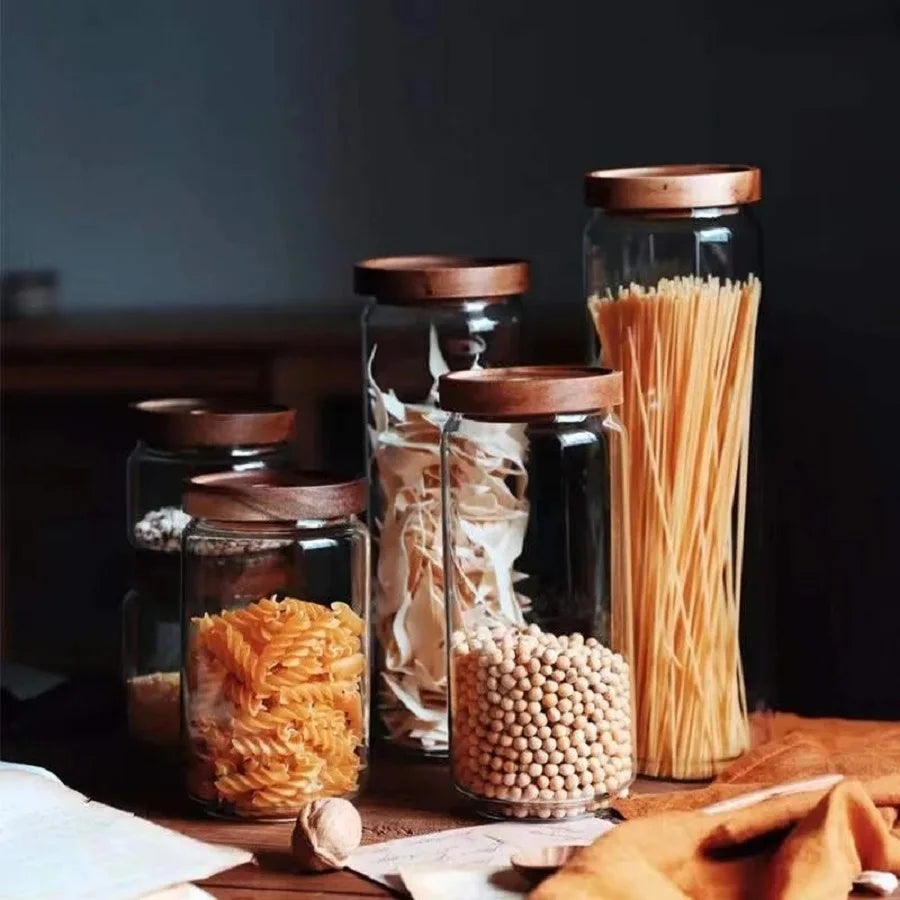 Glass jars with wooden lids containing various types of pasta on a wooden surface.
