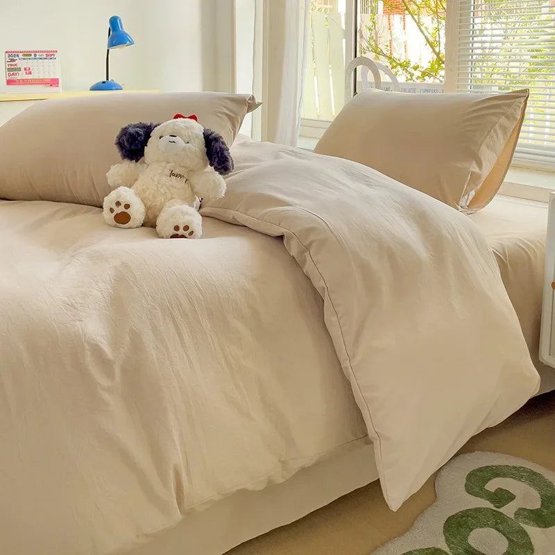 Beige comforter on a bed with a teddy bear, pillows, and a lamp in a bright room.