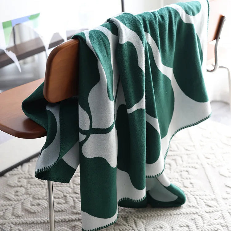 Forest green and white abstract jacquard blanket with blanket stitch edge draped over a wooden chair on a textured cream rug