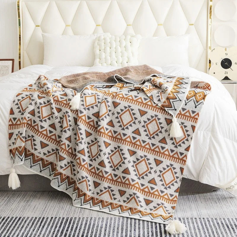 A woven throw blanket with a geometric Southwestern pattern in brown, beige, and white, draped over a bed in a warm, sunlit bedroom.