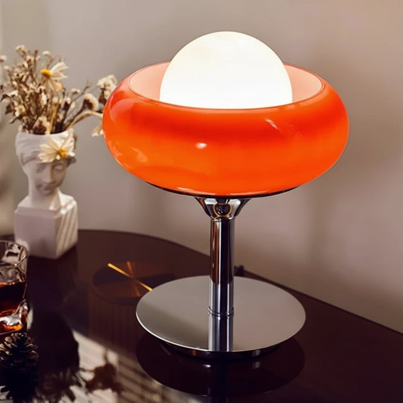 Table lamp with orange base and white shade on a table with a glass of drink and vase.