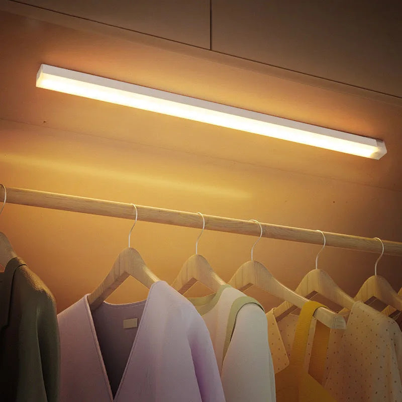 Under-cabinet light fixture illuminating a row of hanging clothes in a closet.