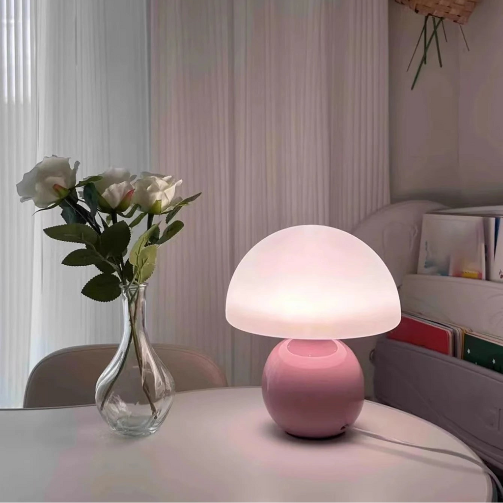 Pink mushroom-shaped lamp on a table with a vase of flowers and a curtain in the background