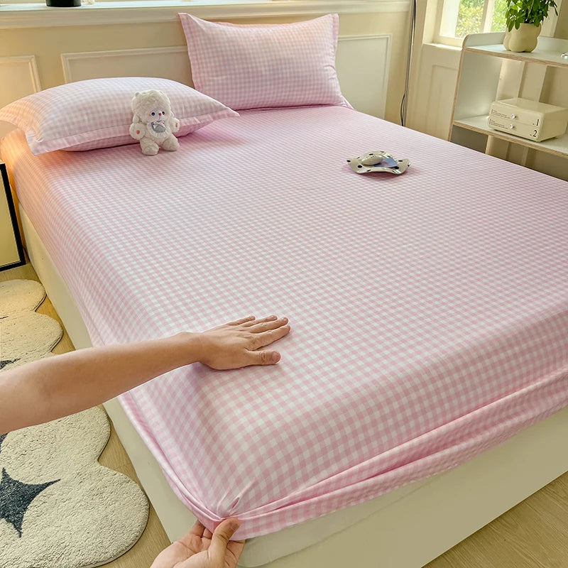 A close-up view of a hand smoothing a soft pink and white plaid fitted sheet onto a mattress. The room features warm lighting and minimalist decor.