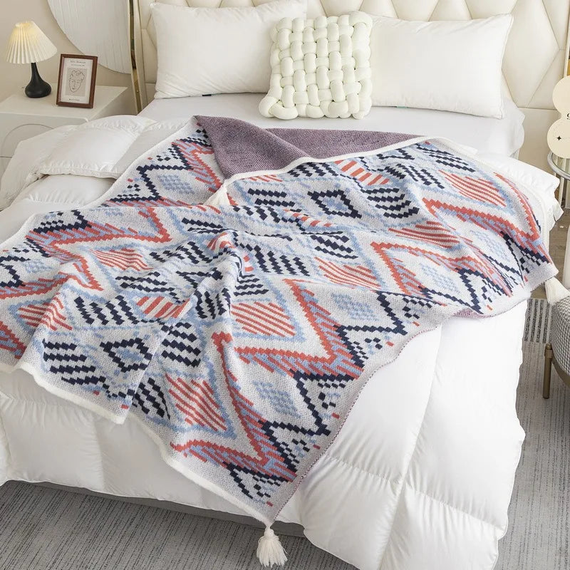 A patterned throw blanket draped over a bed in a bright, modern bedroom. The blanket features a geometric Southwestern-style design in navy blue, terracotta orange, and cream, with a solid cream border and a soft mauve-pink reverse side.