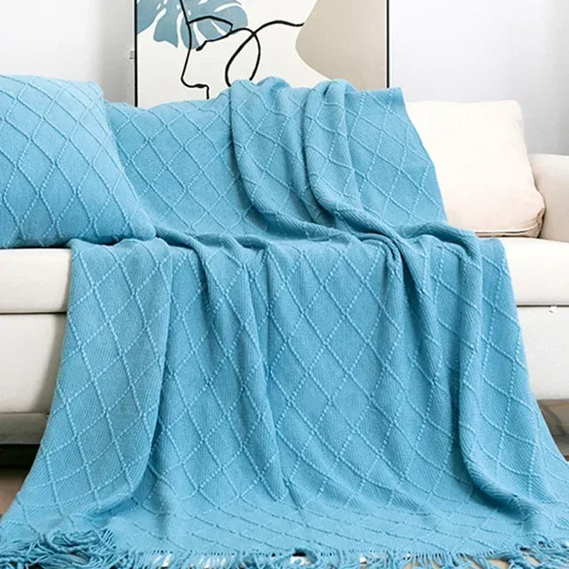 Teal blue diamond-textured knit throw with tassel fringe draped over a cream sofa beside a matching cushion