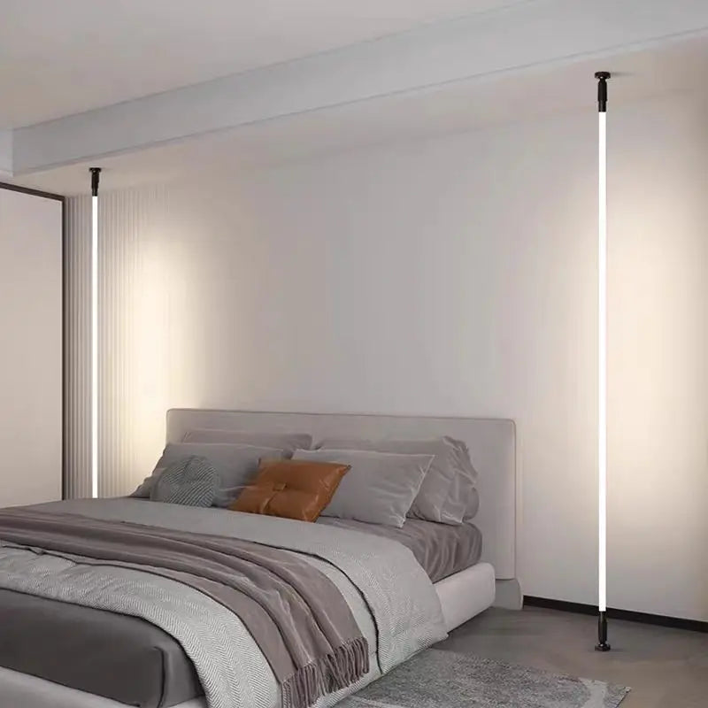 A modern upholstered bed in light gray fabric with grey and brown pillows, set in a minimalist bedroom with two vertical floor lamps on either side of a beige wall.