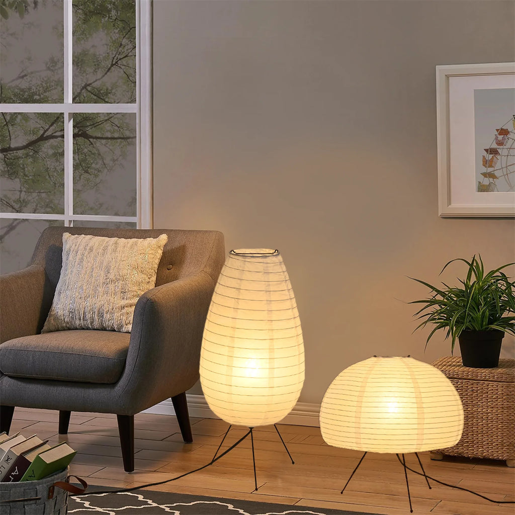 Two illuminated paper lamps on a wooden floor with a chair and plant in the background.